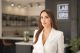 Professional woman in a white blazer standing in a modern office environment with a blurred background and a LAB sign behind her.