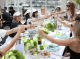 Group of friends toasting with wine glasses around a long table outdoors by a marina harbor.