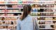 Person with a blue denim jacket and brown strap bag stands in a grocery store aisle facing shelves filled with packaged goods.
