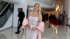 Smiling woman in a silver beaded gown with pink cape sleeves, holding a glittery clutch at a formal indoor event with a red carpet in the background.