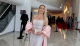 Smiling woman in a silver beaded gown with pink cape sleeves, holding a glittery clutch at a formal indoor event with a red carpet in the background.