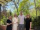 Four adults pose together on a wooden patio with green trees and string lights overhead, outdoors on a sunny day.