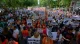 Large diverse crowd of protesters in a sunny park, holding signs demanding housing and rights in Spanish (e.g., 'Derecho a techo', 'Sin casa? a la calle').