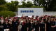 Group of protesters wearing black shirts and red headbands raising fists in front of a Samsung sign in an outdoor area.