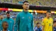 Male footballer in teal Portugal training jacket stands on the pitch before kickoff with teammates nearby amid a stadium crowd and other players in yellow jerseys nearby, indicating a pre-match moment.