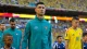 Male footballer in teal Portugal training jacket stands on the pitch before kickoff with teammates nearby amid a stadium crowd and other players in yellow jerseys nearby, indicating a pre-match moment.