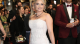 Blonde woman in a beaded off‑white gown posing on a red carpet, clutch in hand, with a man in a burgundy suit behind and a crowd in the background.