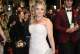 Blonde woman in a beaded off‑white gown posing on a red carpet, clutch in hand, with a man in a burgundy suit behind and a crowd in the background.