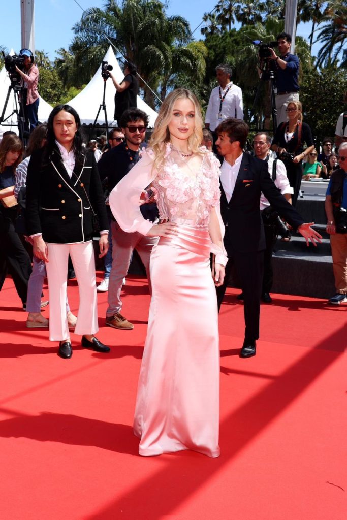 Woman in a pink satin evening gown posing on a red carpet as photographers circle around her outdoors.