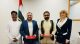 Four people pose in a formal room with the UAE flag in the background; two men hold red folders as if signing an agreement, flanked by a man in white traditional attire and a woman in a navy suit.
