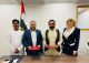 Four people pose in a formal room with the UAE flag in the background; two men hold red folders as if signing an agreement, flanked by a man in white traditional attire and a woman in a navy suit.