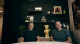 Two men sit at a round white table in a modern room with a dark panel wall, shelves, and a large yellow Pikachu figure between them.