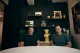 Two men sit at a round white table in a modern room with a dark panel wall, shelves, and a large yellow Pikachu figure between them.