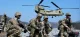 US Army soldiers in camouflage gear kneeling on snowy ground as a CH-47 Chinook helicopter hovers nearby in a mountainous area.