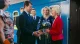 Man in a dark suit shakes hands with a woman in a bright red blazer while a bouquet is handed over, with onlookers and a blue train in the background.