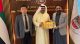 Three men in business attire stand in a formal office lobby with UAE signage and flags; the man in a white kandura holds a row of colorful gift booklets handed to him by the man in a gray blazer.