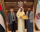 Three men in business attire stand in a formal office lobby with UAE signage and flags; the man in a white kandura holds a row of colorful gift booklets handed to him by the man in a gray blazer.