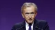 Older man in a dark suit and purple tie speaks at a podium with two microphones against a purple background.
