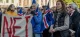 Crowd of protesters in an urban square, waving blue, red, and white flags as a large banner with red letters is held at the front.
