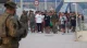 Group of civilians walking along a wide promenade flanked by soldiers in camouflage; a soldier in the foreground is blurred, showing a security presence.