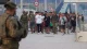Group of civilians walking along a wide promenade flanked by soldiers in camouflage; a soldier in the foreground is blurred, showing a security presence.