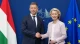 Two formally dressed officials shake hands in front of a blue backdrop with EU and national flags visible (Hungarian flag left).