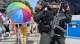 Police officer in full tactical gear with rifle and orange POLIZEI patches stands near a crowd behind metal barriers at a parade or public event, rainbow umbrella in the background suggesting a Pride celebration.