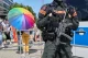 Police officer in full tactical gear with rifle and orange POLIZEI patches stands near a crowd behind metal barriers at a parade or public event, rainbow umbrella in the background suggesting a Pride celebration.