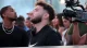 Man with short beard in a black Nike shirt looks to the side as a cameraman films nearby at a crowded event