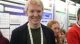 Smiling young man wearing a dark blazer and lanyard at a conference with posters in the background.