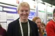 Smiling young man wearing a dark blazer and lanyard at a conference with posters in the background.